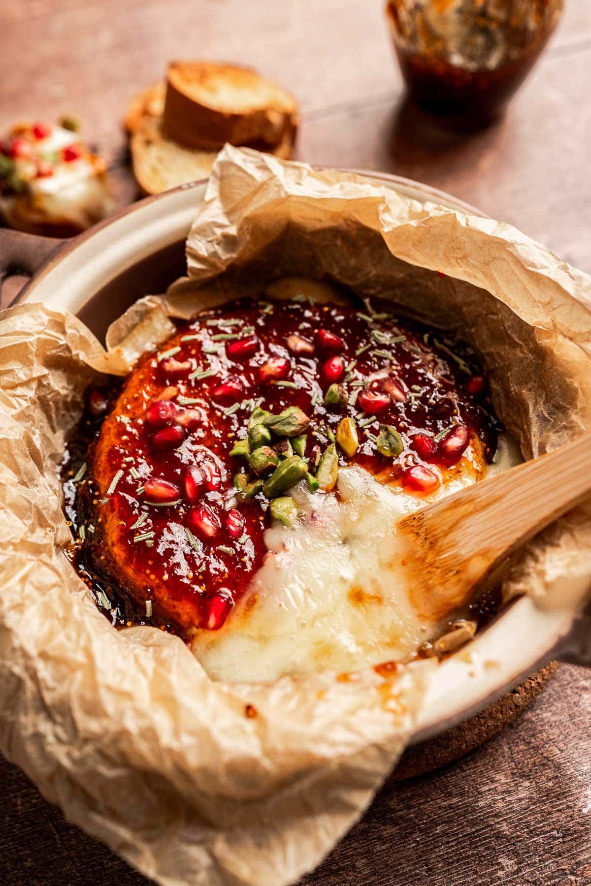 Baked brie and fig jam topped with pomegranate seeds, pistachios, and sauce, served on parchment paper with a wooden spreader and slices of bread in the background.
