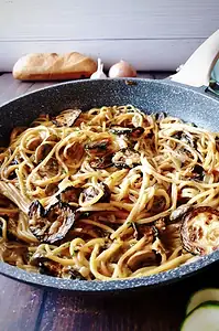 A pan of cooked spaghetti mixed with sliced, roasted zucchini and a creamy sauce, set on a wooden surface with bread and garlic in the background.