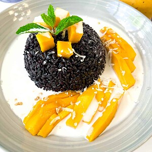 A serving of black sticky rice topped with mango pieces, coconut flakes, and mint, surrounded by coconut sauce and sliced mango on a white plate.