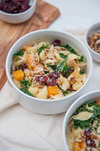 A white bowl filled with pasta shells, kale, roasted butternut squash, dried cranberries, and topped with chopped walnuts and grated cheese.