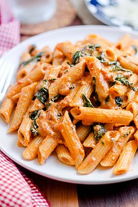 A plate of rigatoni pasta in a creamy tomato sauce with spinach, served on a white dish beside a red and white napkin.