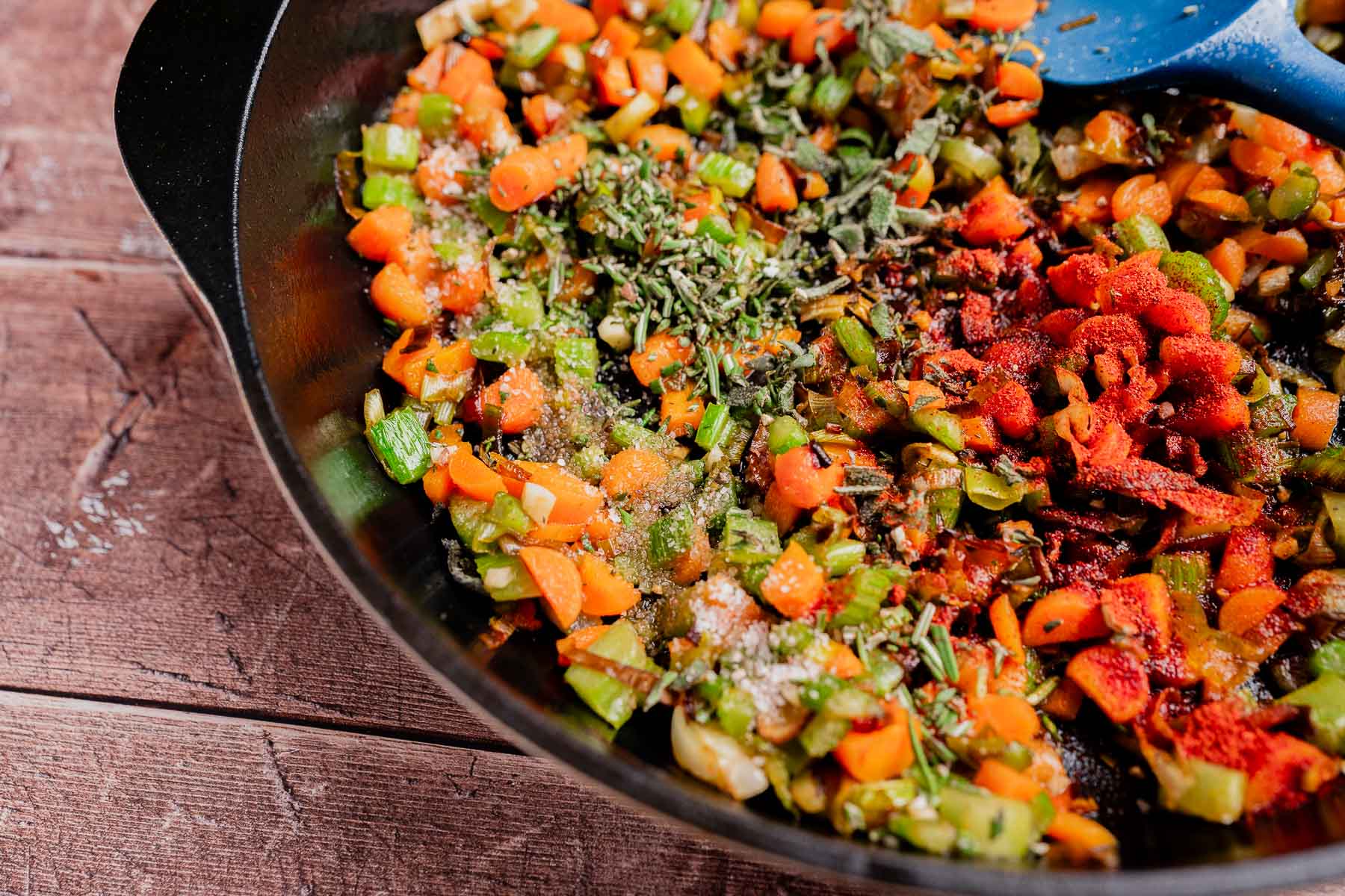 Diced carrots, celery, and green onions with herbs and spices cooking in a black skillet on a wooden surface—perfect as a base for gluten free cornbread stuffing.