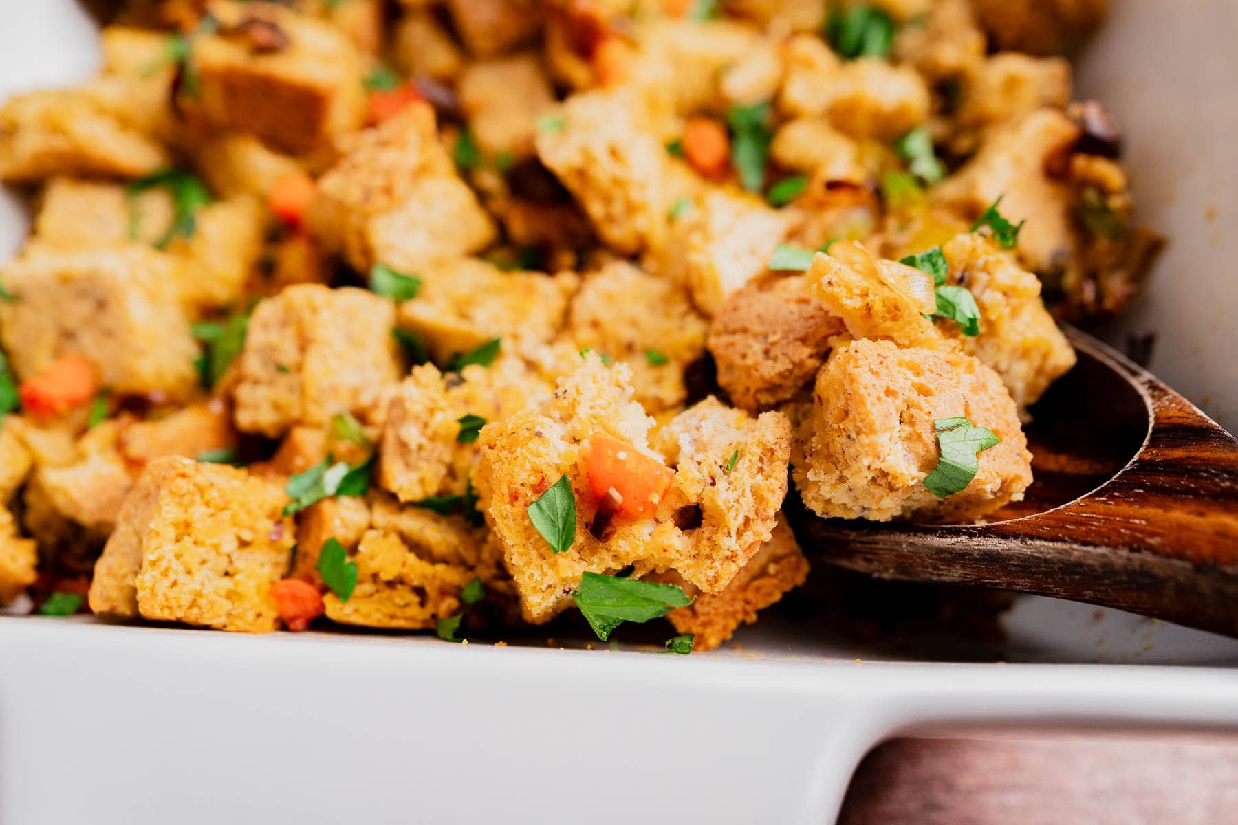 A close-up of gluten free cornbread stuffing with herbs and vegetables in a white dish, being served with a wooden spoon.