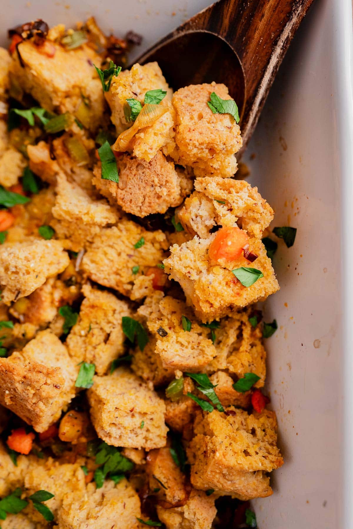Close-up of gluten free cornbread stuffing with chopped vegetables and herbs in a white dish, with a wooden spoon scooping some pieces.