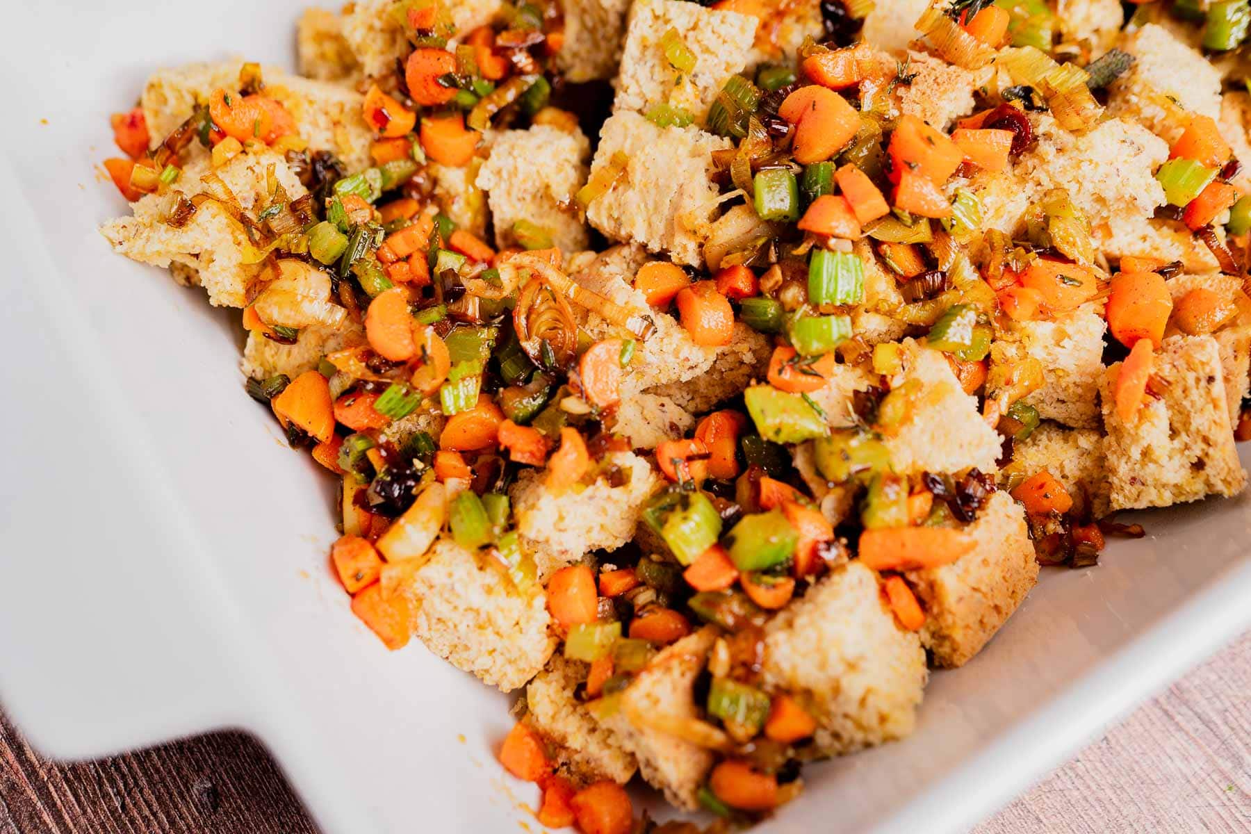 A close-up of gluten free cornbread stuffing in a white dish, showing cubed bread mixed with diced carrots, celery, and herbs.