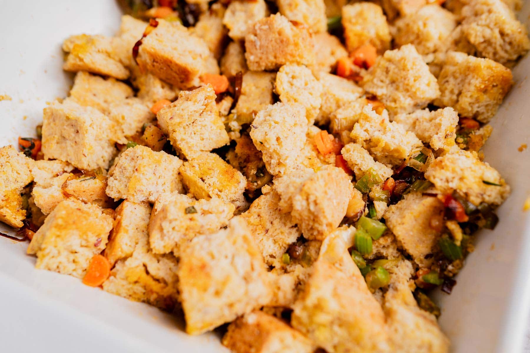 Close-up of homemade gluten free cornbread stuffing with cubed bread, chopped vegetables, and savory seasoning in a white dish.