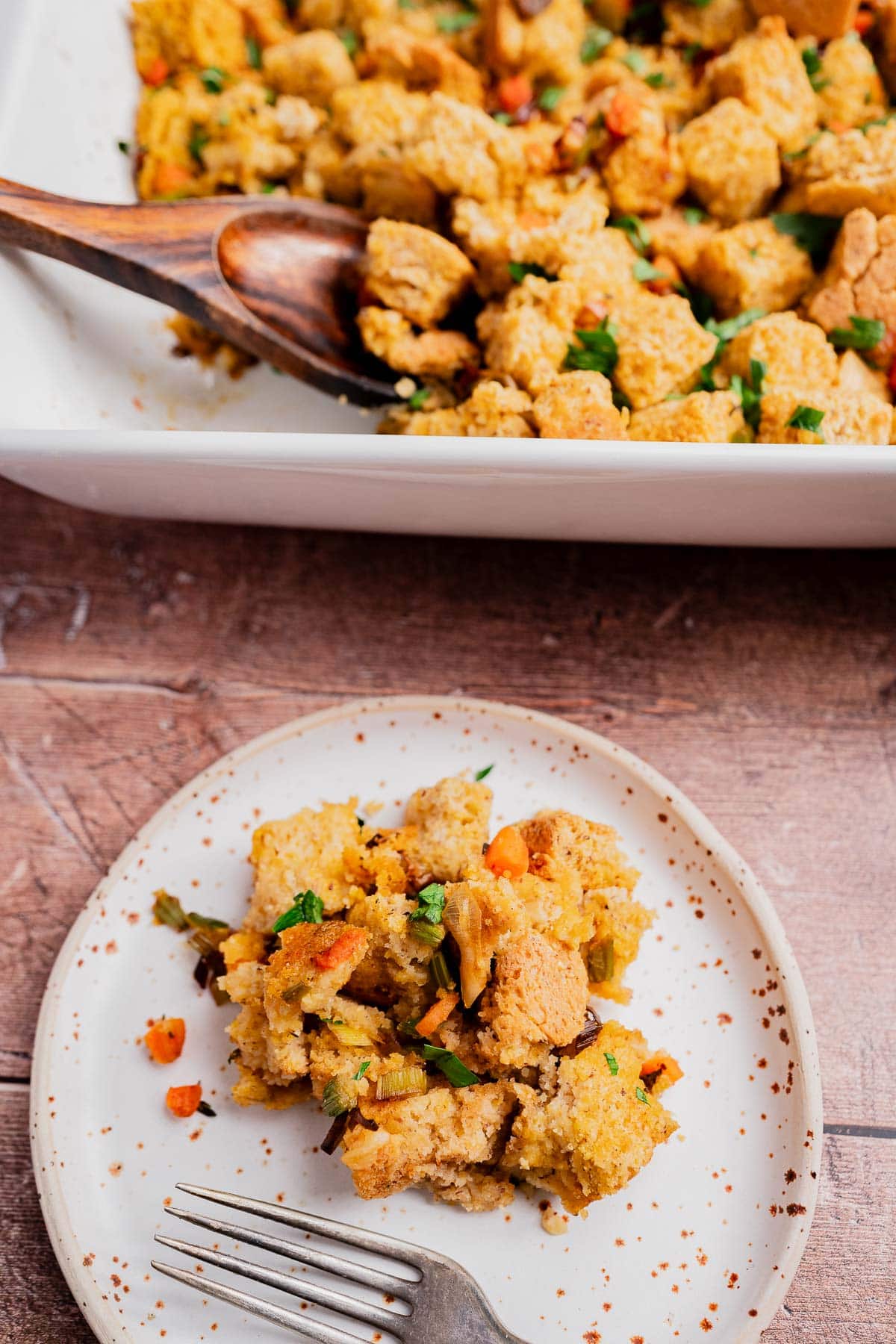 A plate with a serving of gluten free cornbread stuffing, featuring visible vegetables, sits in front of a baking dish with more stuffing and a wooden spoon.