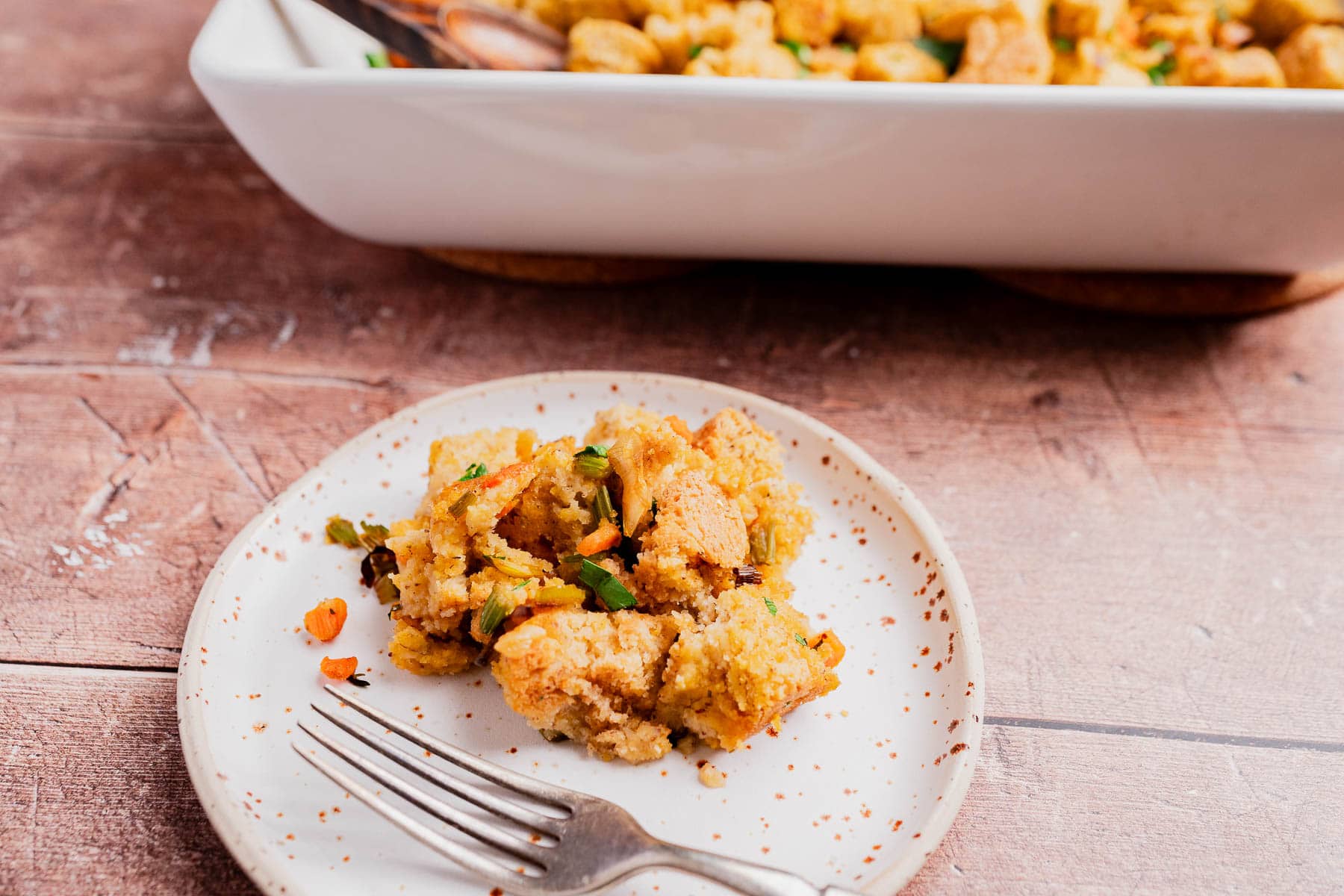 A serving of gluten free cornbread stuffing with vegetables on a white speckled plate, a fork beside it, and a baking dish of stuffing in the background.