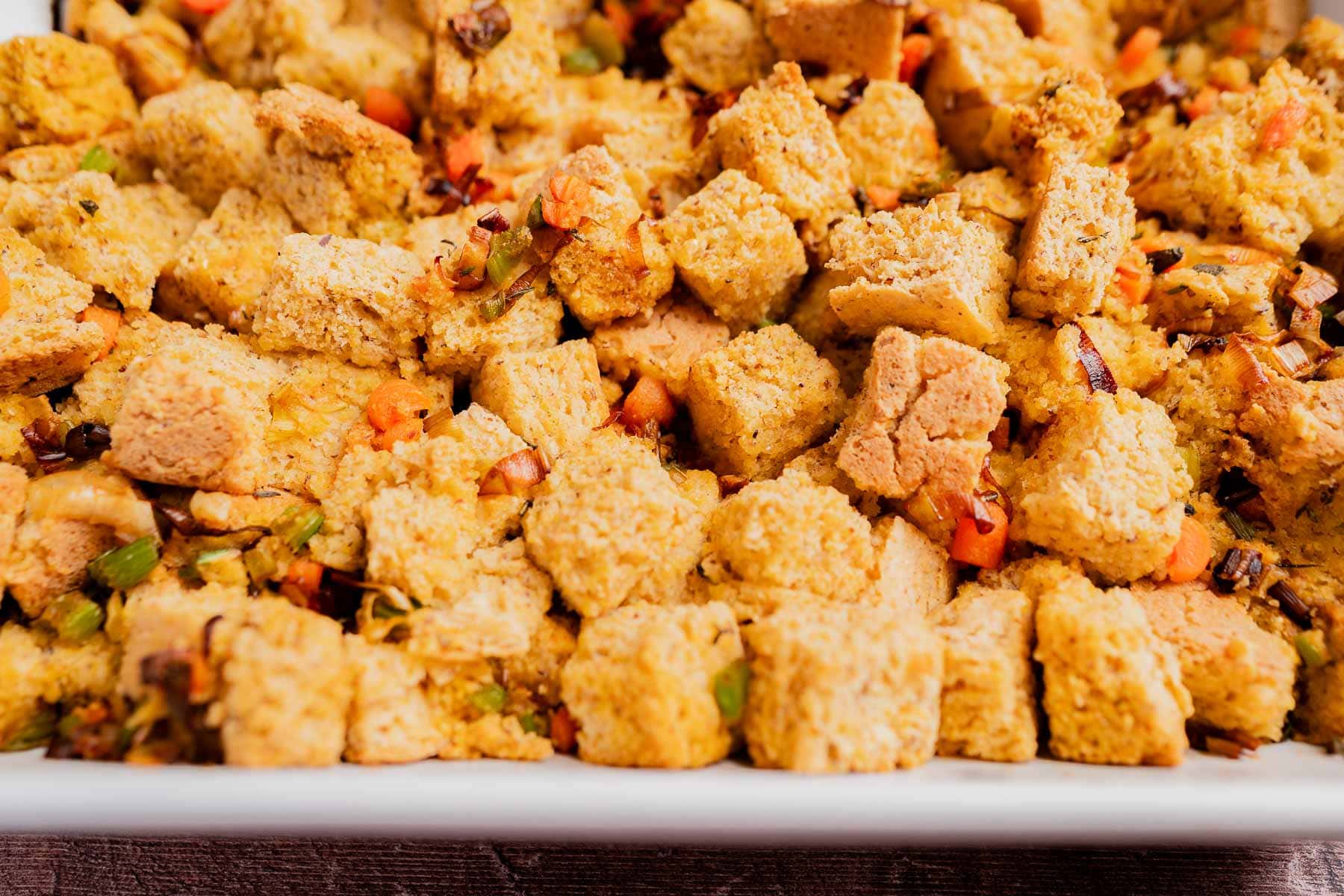 A close-up of gluten free cornbread stuffing with toasted bread cubes, diced vegetables, and seasonings in a white dish.