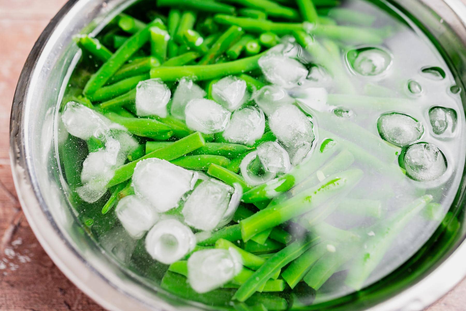 Green beans in a metal bowl filled with water and ice cubes, being chilled in an ice bath—the perfect prep step for a gluten free vegan green bean casserole.