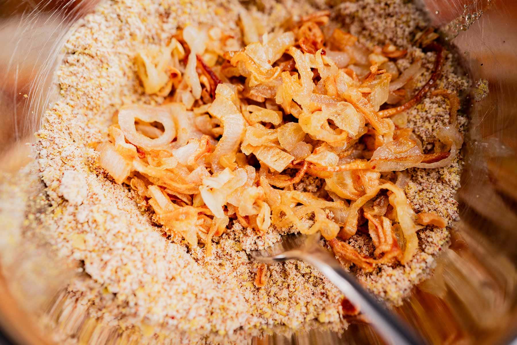 Sliced, sautéed onions rest atop a mixture of dry breadcrumbs and seasonings in a metal bowl with a spoon, perfect for preparing a gluten free vegan green bean casserole.