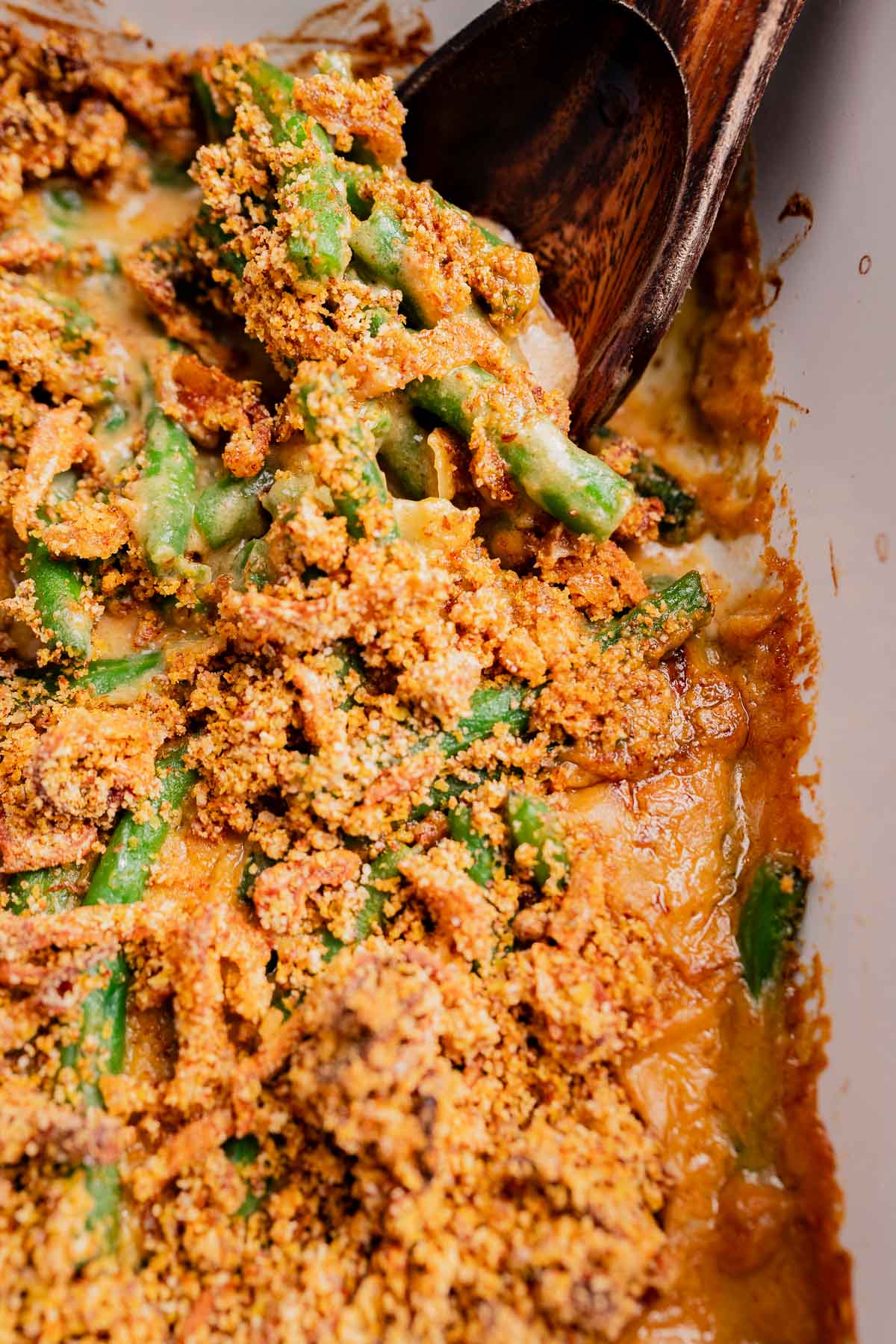 Close-up of gluten free vegan green bean casserole with a crispy, golden breadcrumb topping in a baking dish, being served with a wooden spoon.