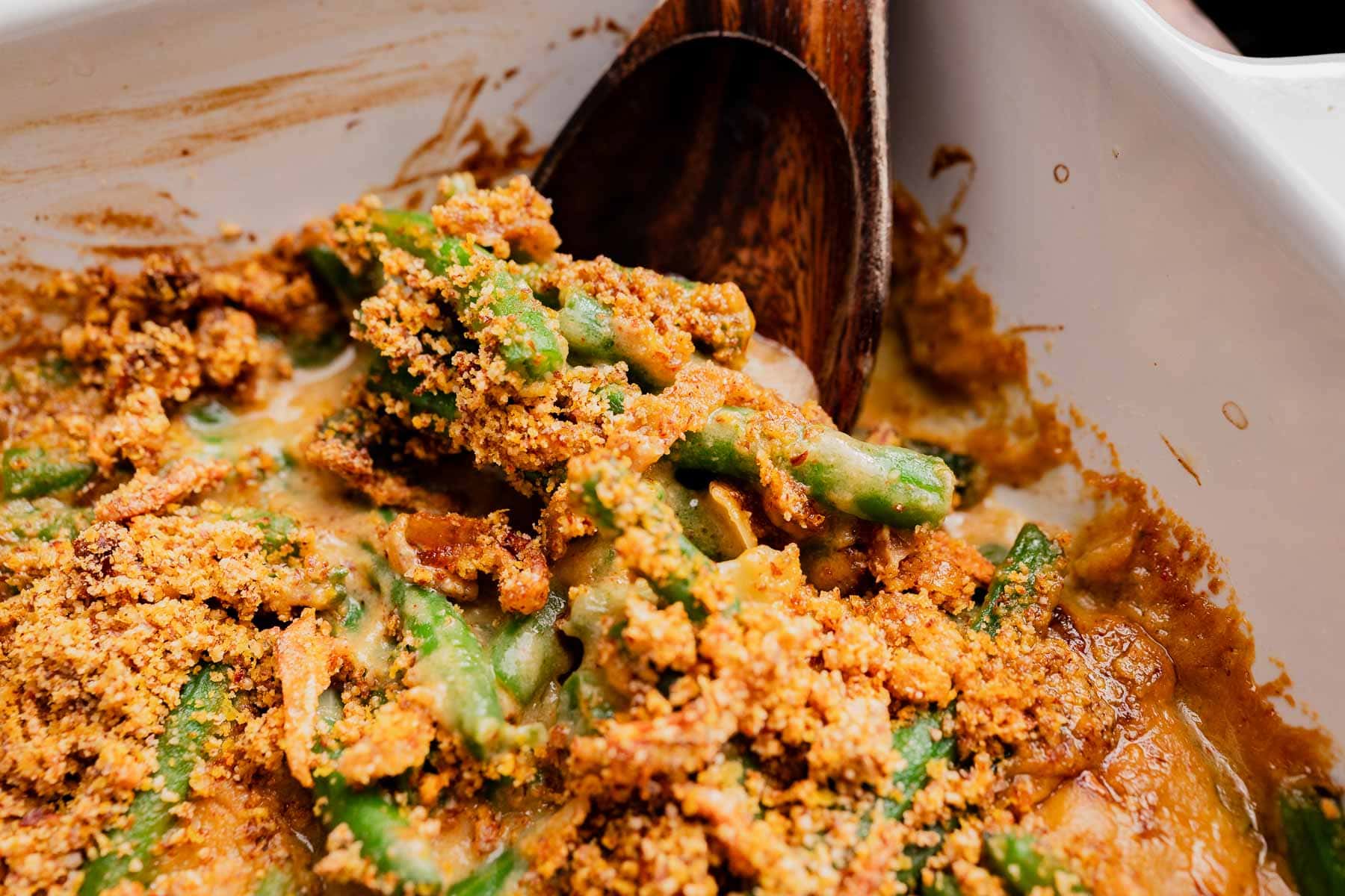 Close-up of a gluten free vegan green bean casserole topped with crispy breadcrumbs in a white baking dish, with a wooden serving spoon.