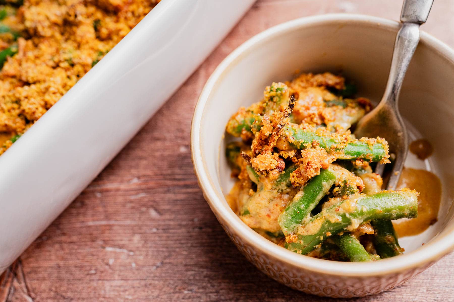A bowl of gluten free vegan green bean casserole topped with crispy breadcrumbs sits next to a baking dish on a wooden surface, with a spoon in the bowl.