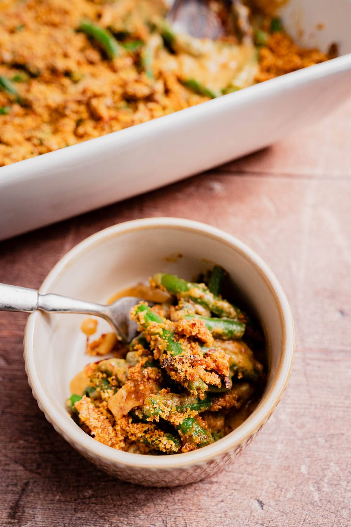 A bowl of gluten free vegan green bean casserole with a crispy breadcrumb topping and a fork, next to a baking dish with more casserole on a wooden surface.