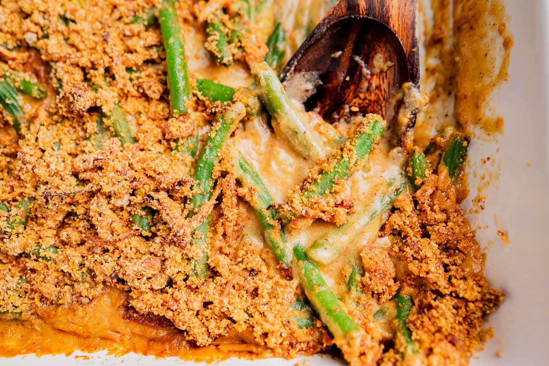 Close-up of a gluten free vegan green bean casserole with a creamy sauce and breadcrumb topping, partially scooped with a wooden spoon.