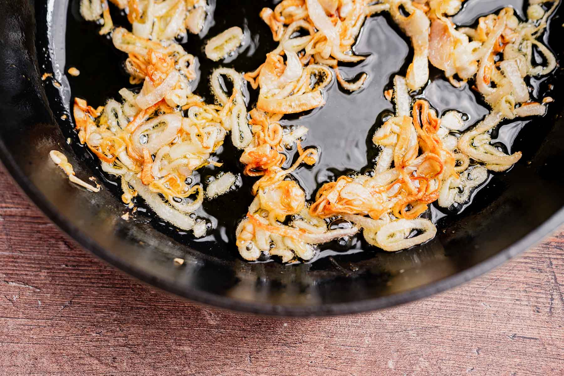 Thinly sliced onions frying in oil in a black skillet, with some pieces golden brown and crispy—a perfect topping for your gluten free vegan green bean casserole.