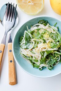 A bowl of creamy spinach and noodle salad is placed next to a halved lemon and wooden-handled fork and spoon on a white surface.