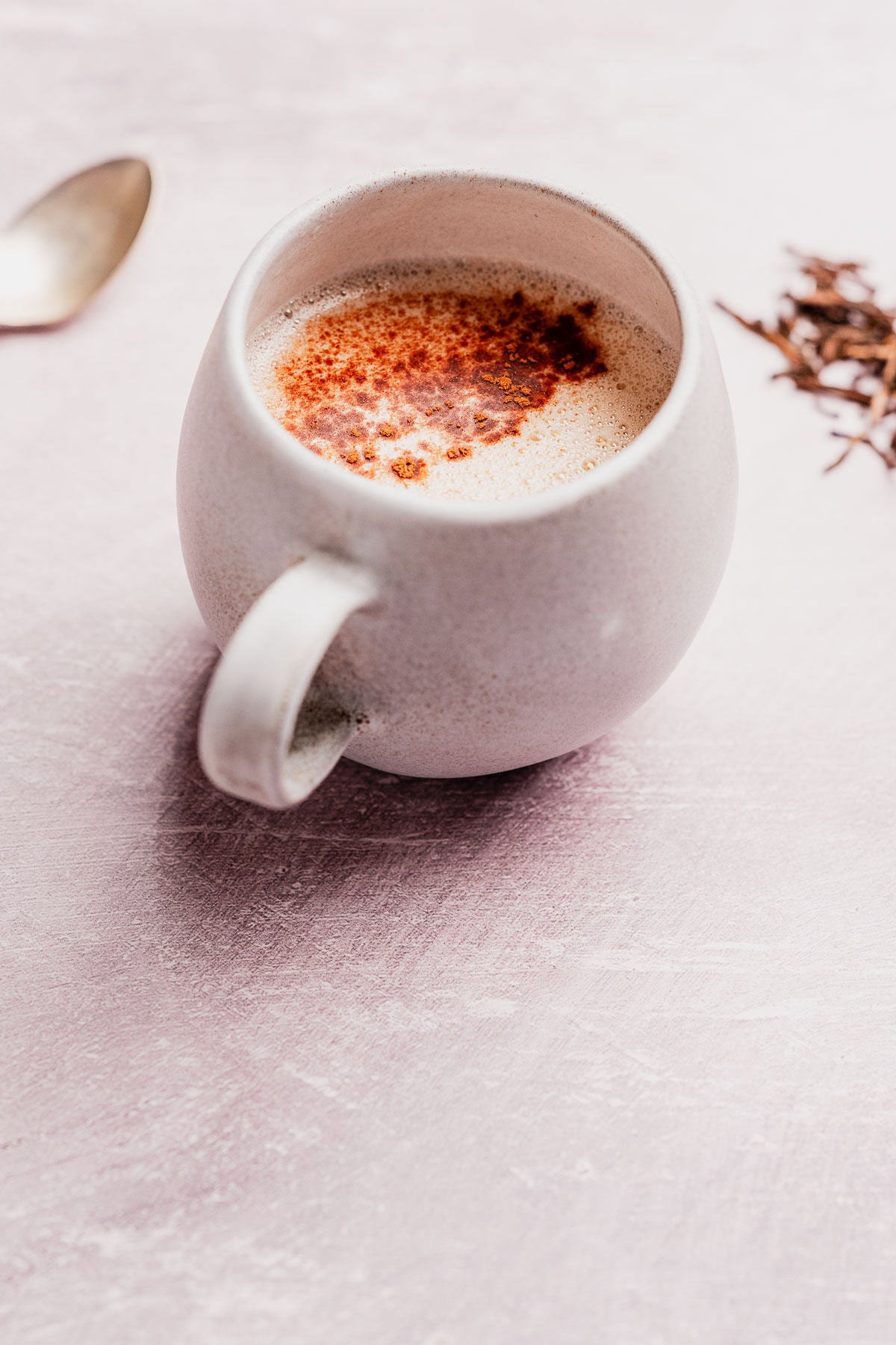 A white ceramic mug filled with a frothy hojicha latte, sprinkled with cinnamon, sits on a light pink surface next to a spoon and some loose tea leaves.