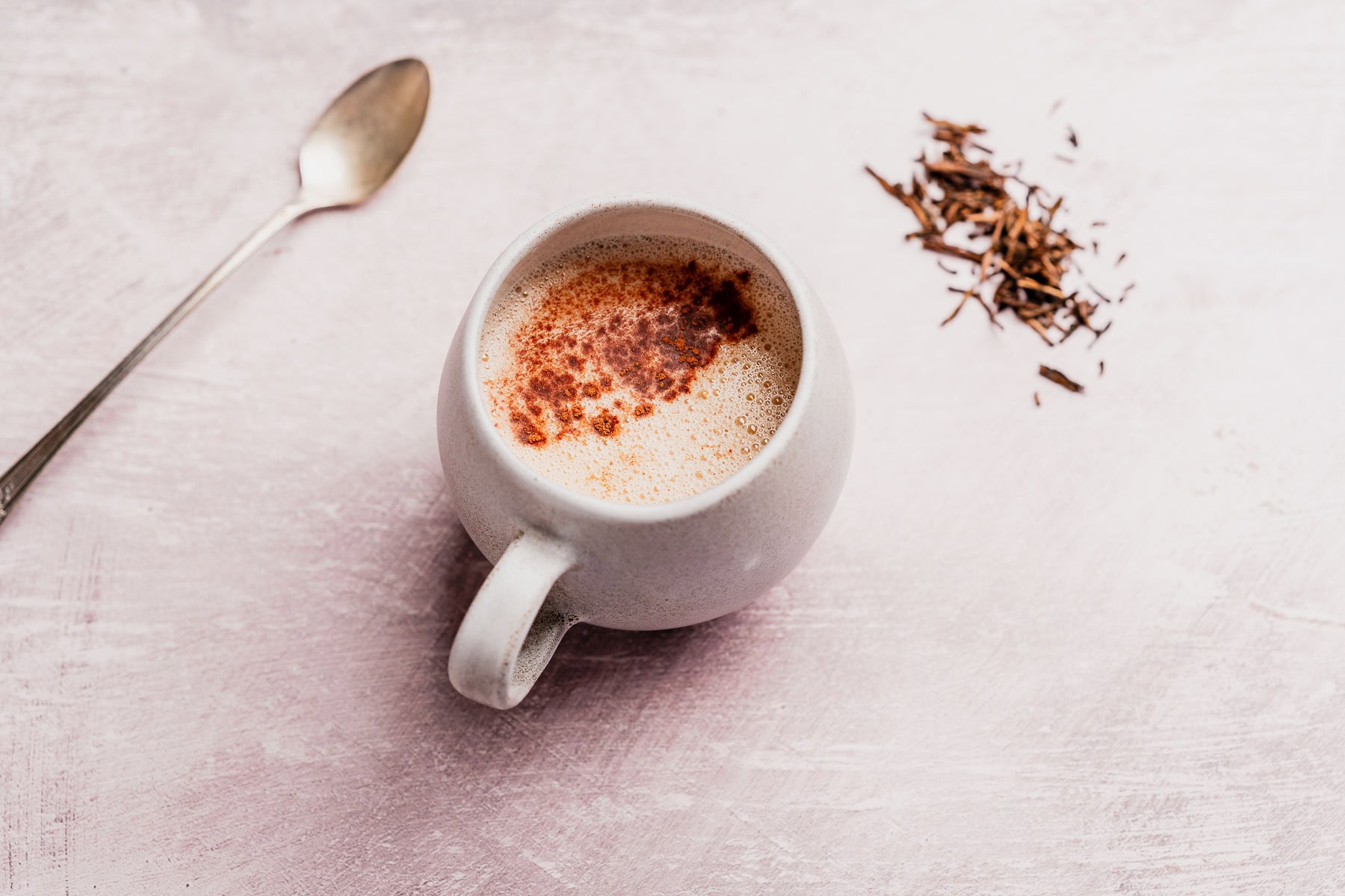 A white mug filled with a frothy hojicha latte topped with cinnamon sits on a light pink surface next to a spoon and a small pile of loose tea leaves.