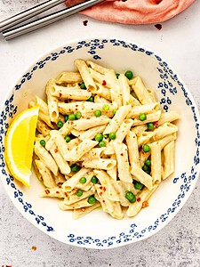 A bowl of penne pasta with green peas, creamy sauce, red pepper flakes, and a lemon wedge, served in a patterned white dish.