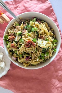 A white bowl of orzo pasta salad with feta, sun-dried tomatoes, green peas, red onion, and greens sits on a pink cloth with a fork in the bowl.