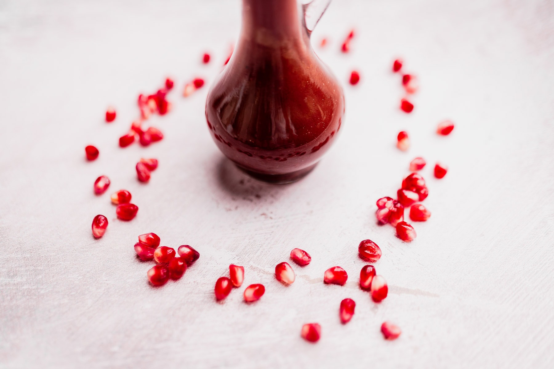A glass bottle filled with dark red Pomegranate Salad Dressing is surrounded by scattered pomegranate seeds on a light surface.