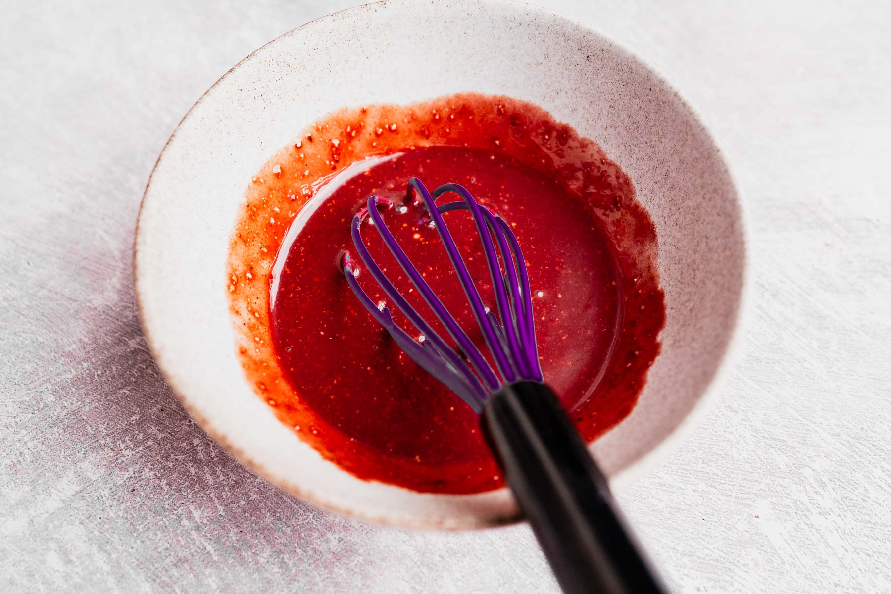 A white bowl containing a vibrant red Pomegranate Salad Dressing mixture and a purple whisk rests on a light-colored surface.