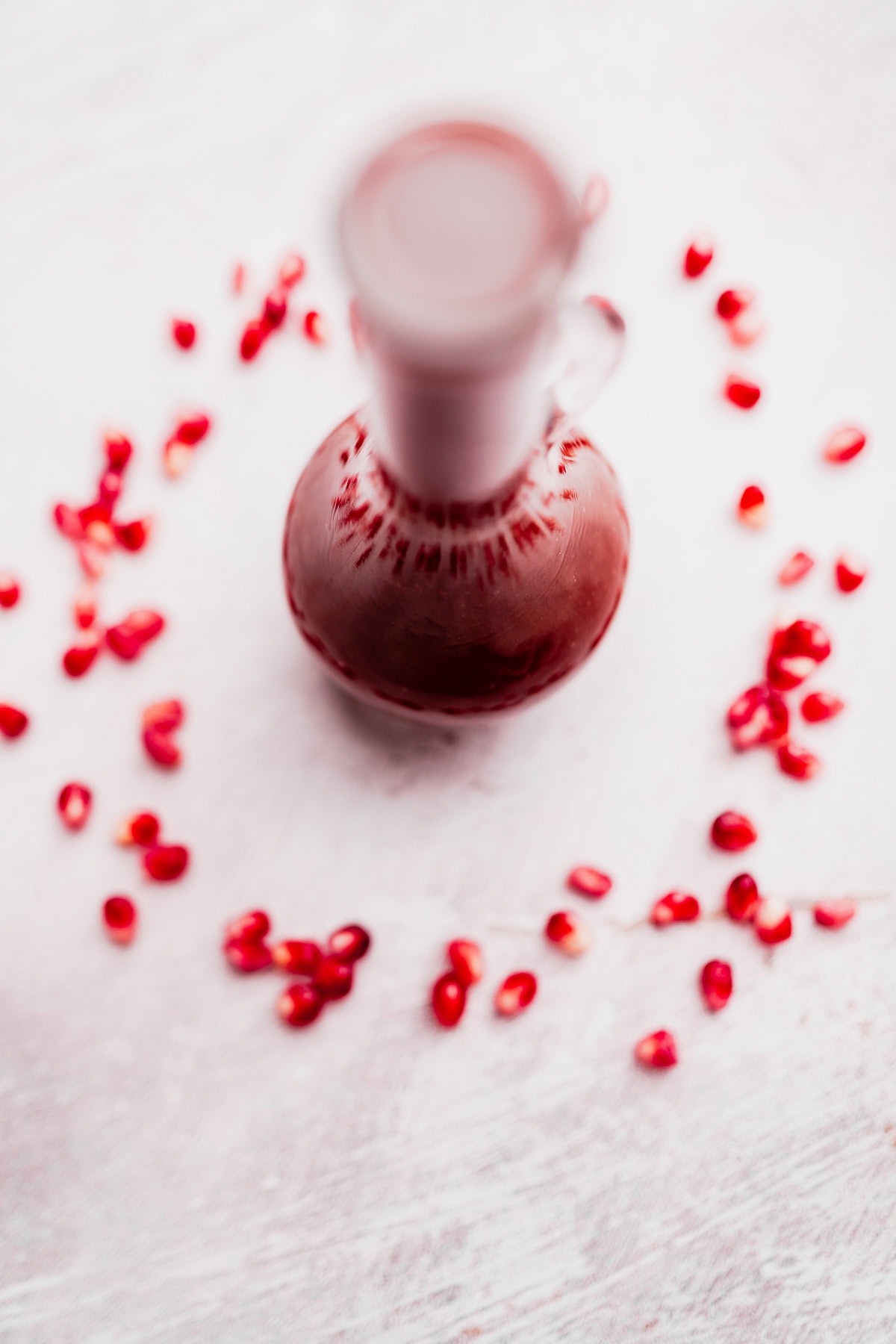 A glass jug filled with red liquid is placed on a white surface, surrounded by scattered pomegranate seeds, suggesting it holds vibrant Pomegranate Salad Dressing.