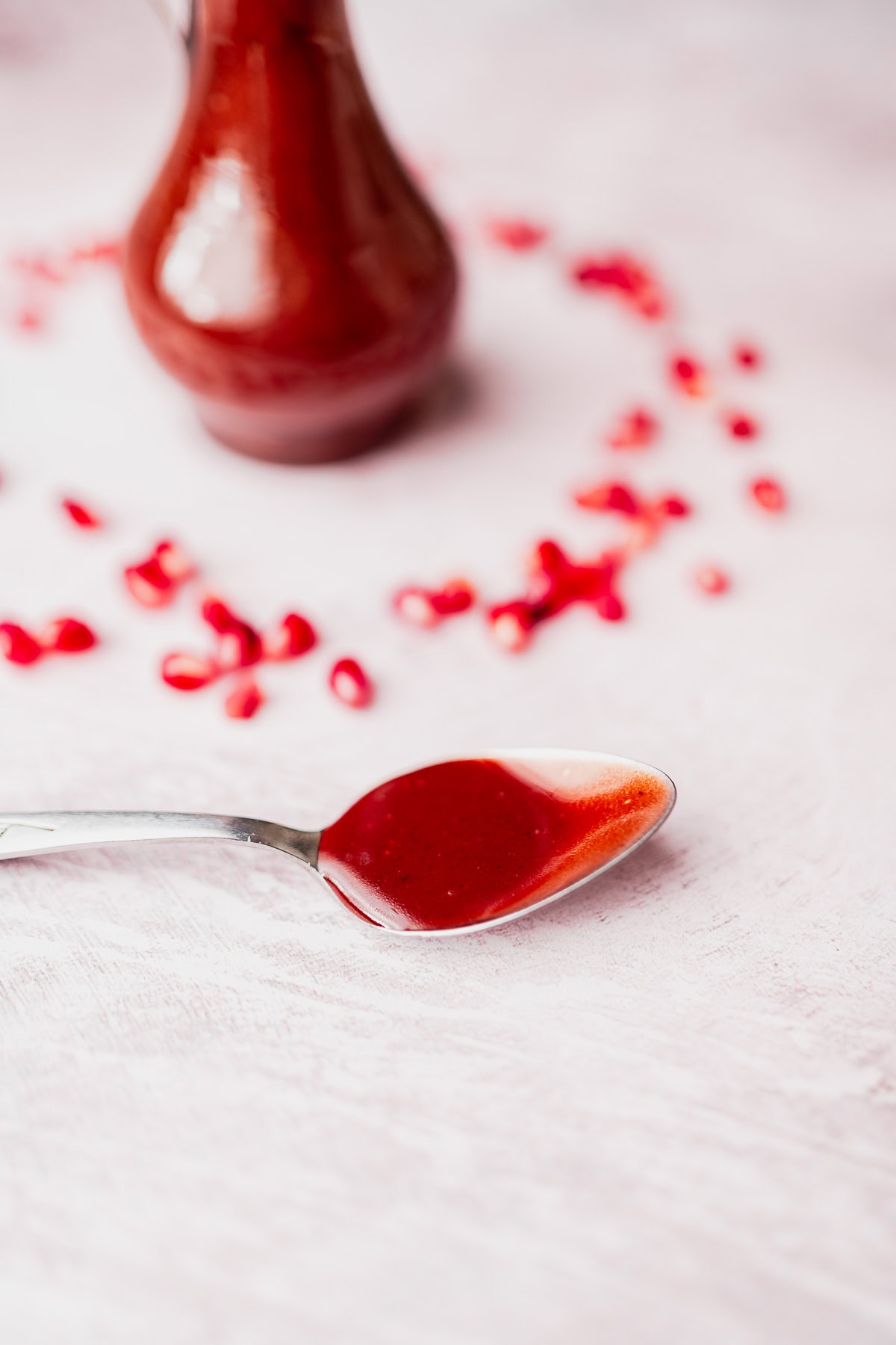 A spoon with vibrant red Pomegranate Salad Dressing in the foreground, with a glass bottle of sauce and scattered pomegranate seeds on a light pink surface in the background.