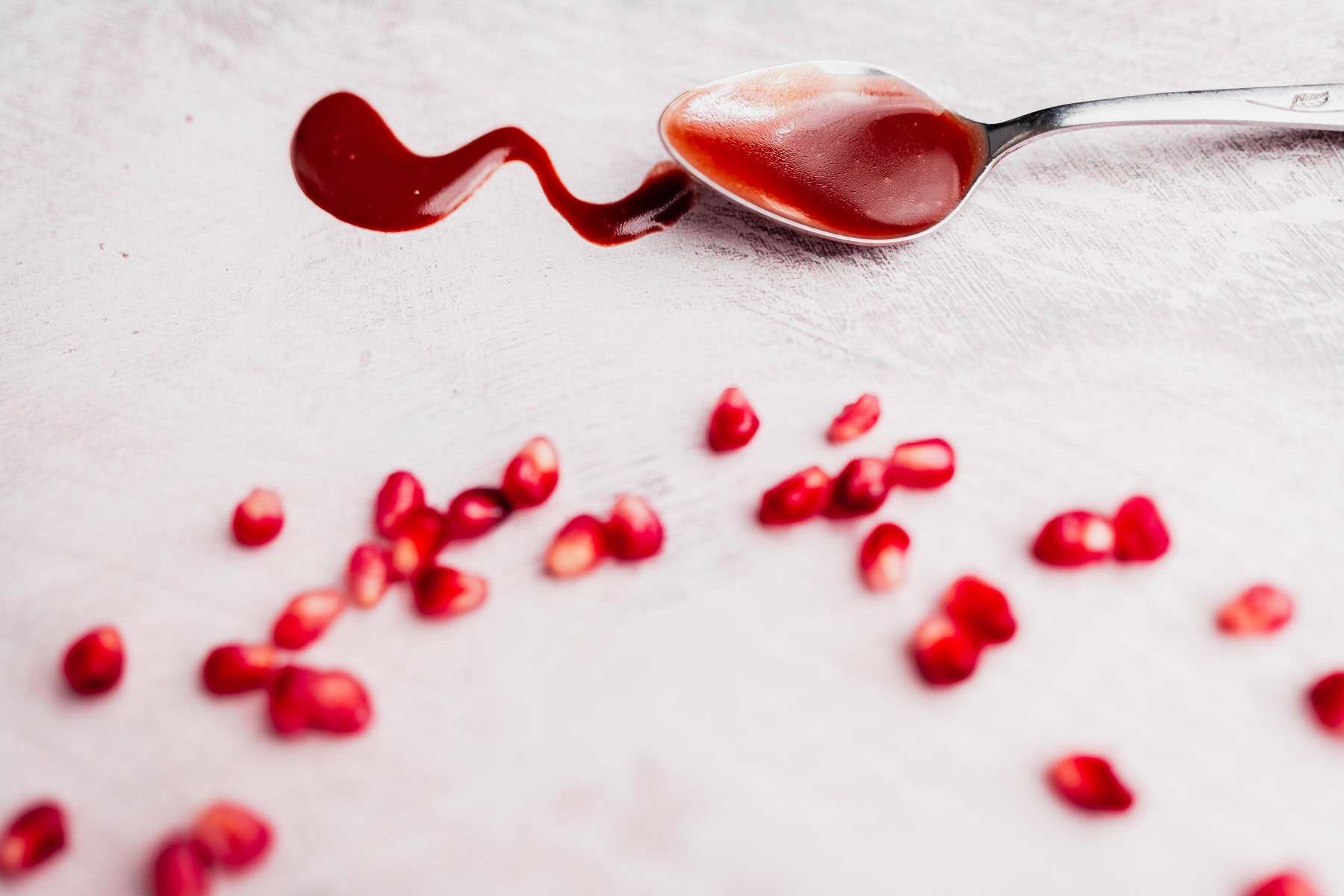 A spoon with pomegranate juice and scattered pomegranate seeds on a light textured surface, perfect for making vibrant Pomegranate Salad Dressing.