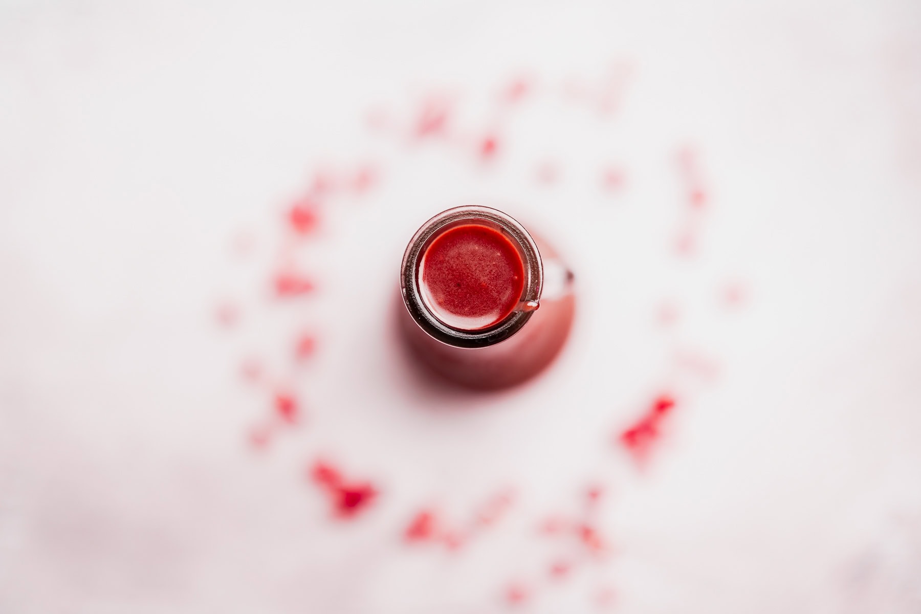 A top-down view of a glass bottle filled with vibrant red pomegranate salad dressing, surrounded by scattered red petals or droplets on a white surface.