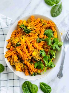 A bowl of rotini pasta with vegetables and sauce, garnished with cilantro, on a marble surface with spinach leaves and a fork nearby.