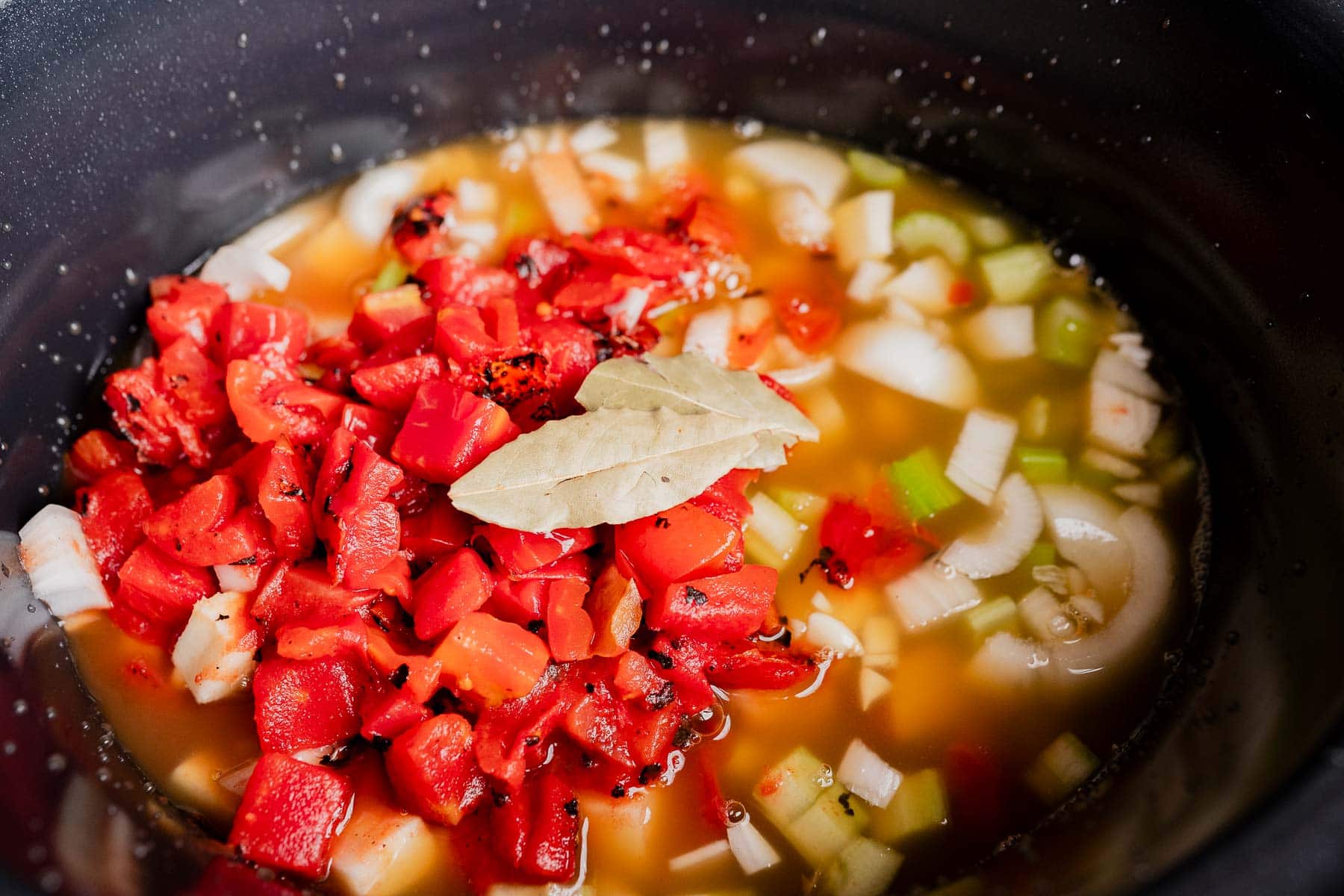 Chopped tomatoes, onions, celery, and a bay leaf in broth inside a pot, ready to become a delicious slow cooker vegetable stew.