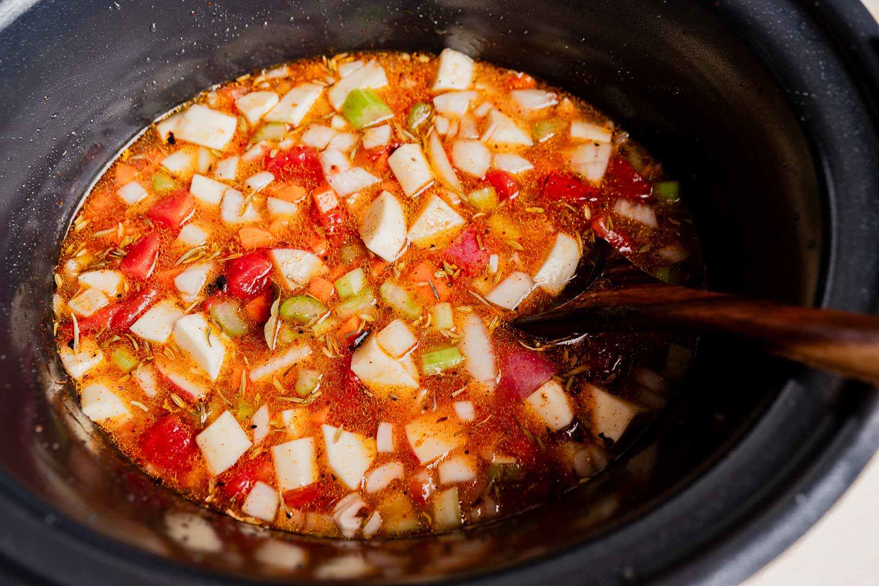Chopped vegetables and broth in a slow cooker, with a wooden spoon, ready to be cooked into a hearty slow cooker vegetable stew or soup.