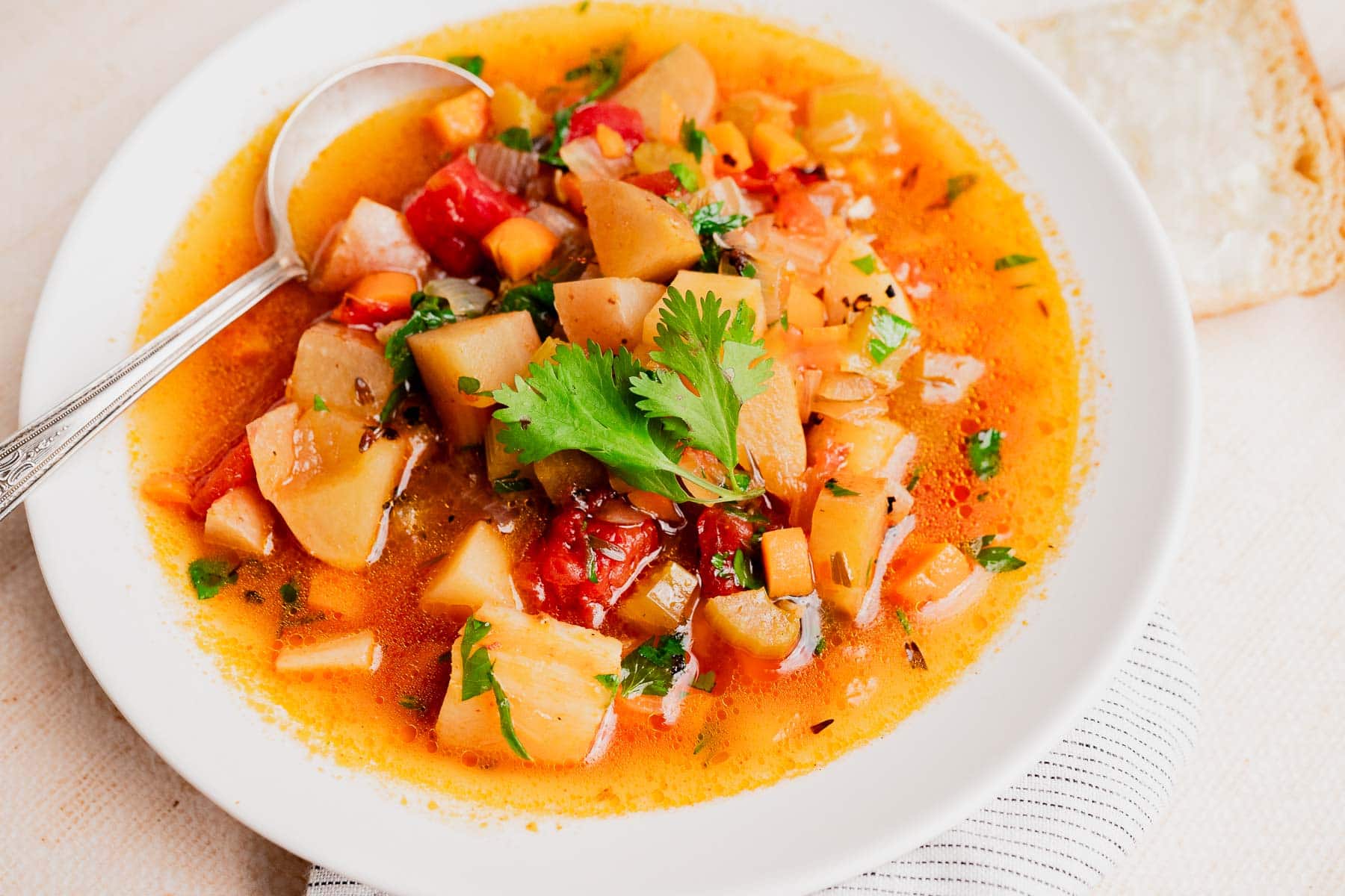 A bowl of slow cooker vegetable stew with potatoes, carrots, tomatoes, and fresh cilantro garnish, served with a spoon and a slice of bread on the side.