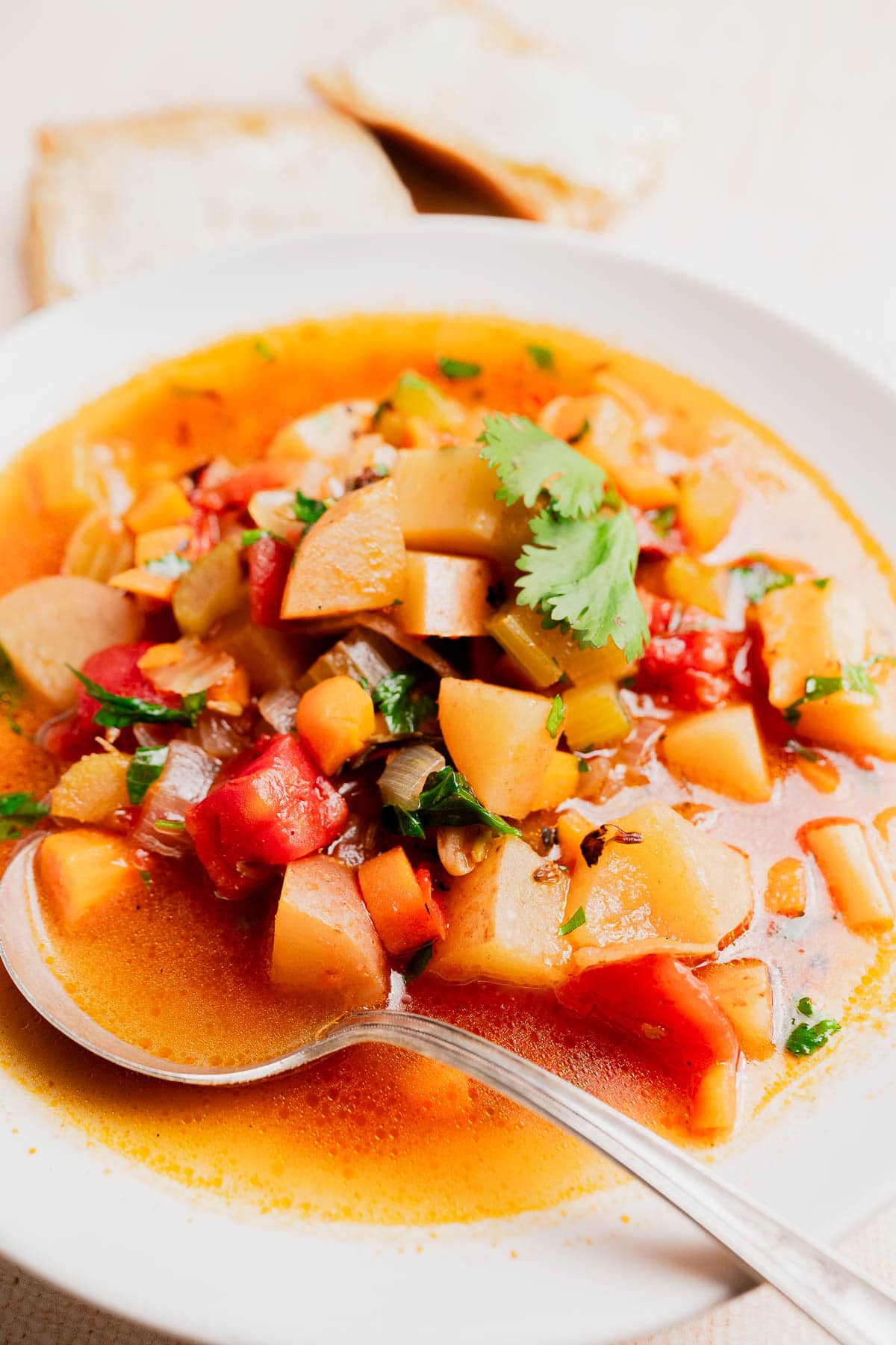 A bowl of slow cooker vegetable stew with potatoes, carrots, and tomatoes, garnished with cilantro, with a spoon in the bowl and bread slices in the background.
