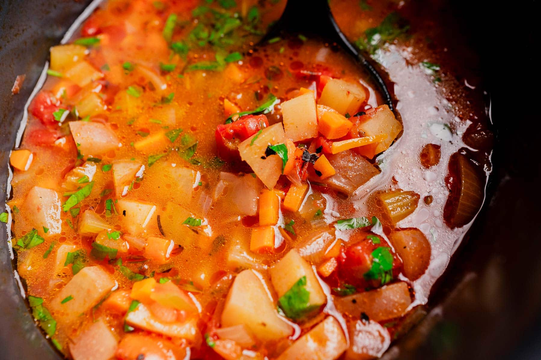 A close-up of slow cooker vegetable stew with diced potatoes, carrots, tomatoes, and fresh herbs in a broth, being stirred with a black ladle.