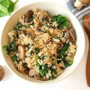 A bowl of rice mixed with sautéed mushrooms, spinach, and onions, placed on a white surface with a spinach leaf and mushroom nearby.