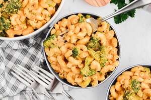 A bowl of macaroni and cheese mixed with broccoli, placed on a checkered cloth with additional bowls and forks nearby.