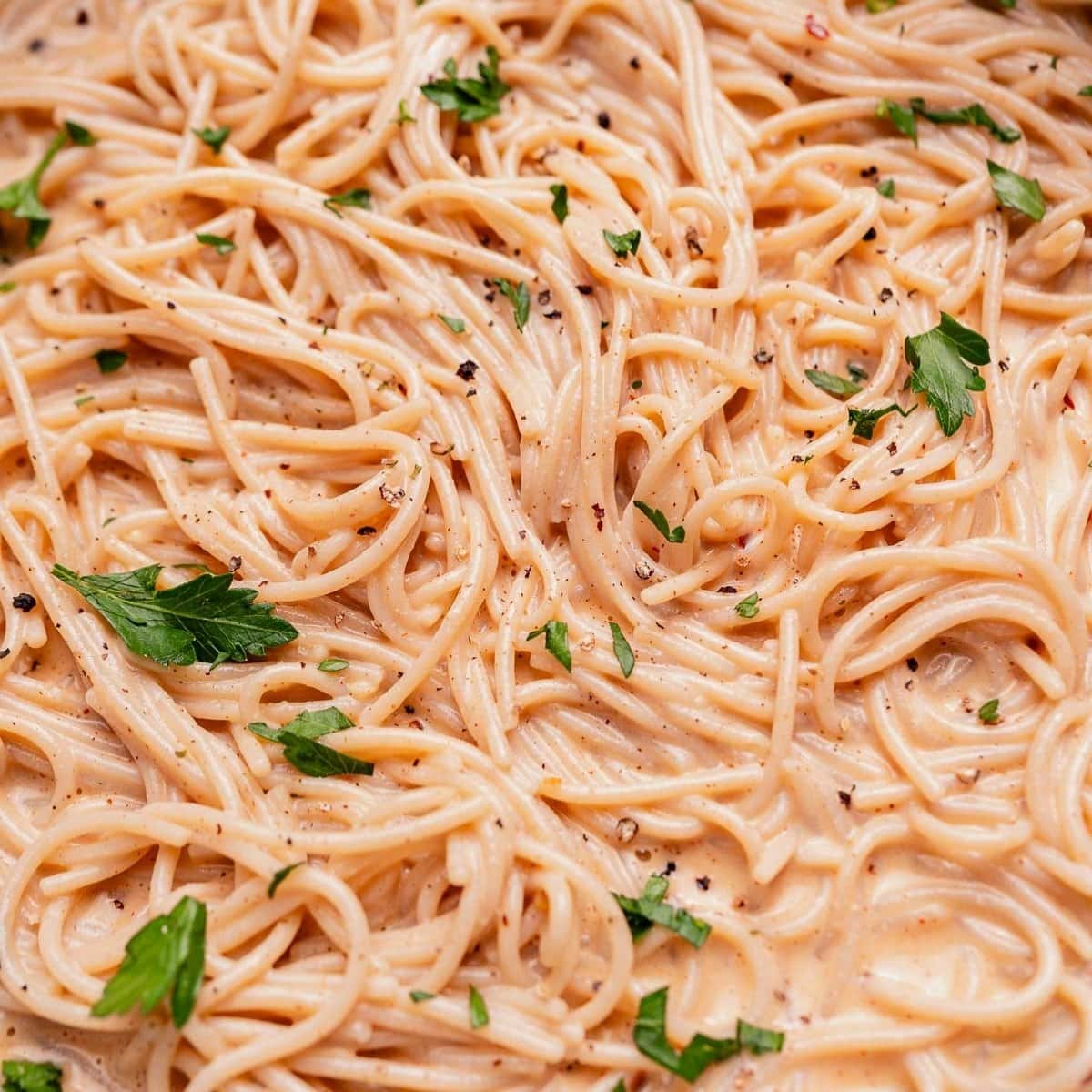 Close-up of cooked spaghetti in a creamy sauce, garnished with chopped parsley and cracked black pepper.