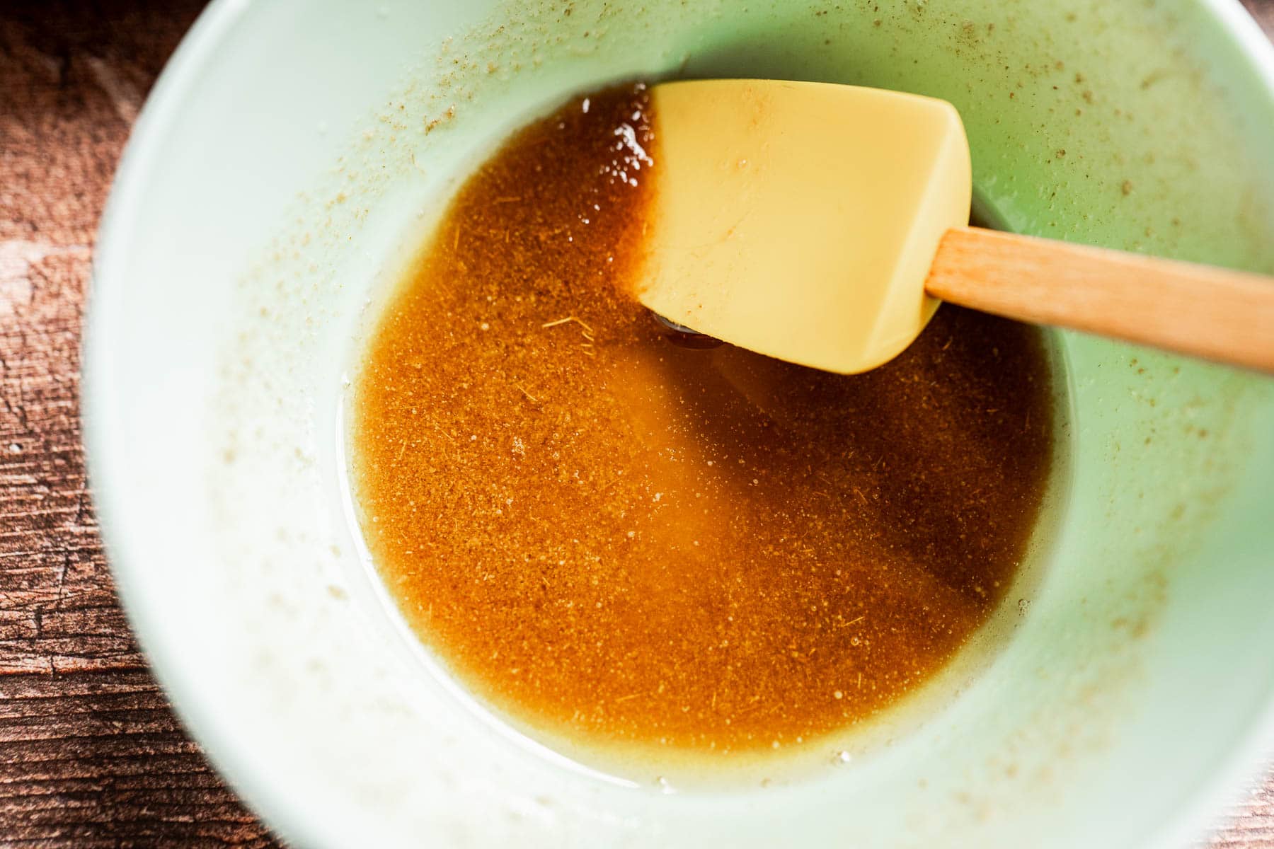 A mixing bowl containing a brown liquid mixture—perfect for making brown sugar glazed carrots—with a yellow spatula resting inside.