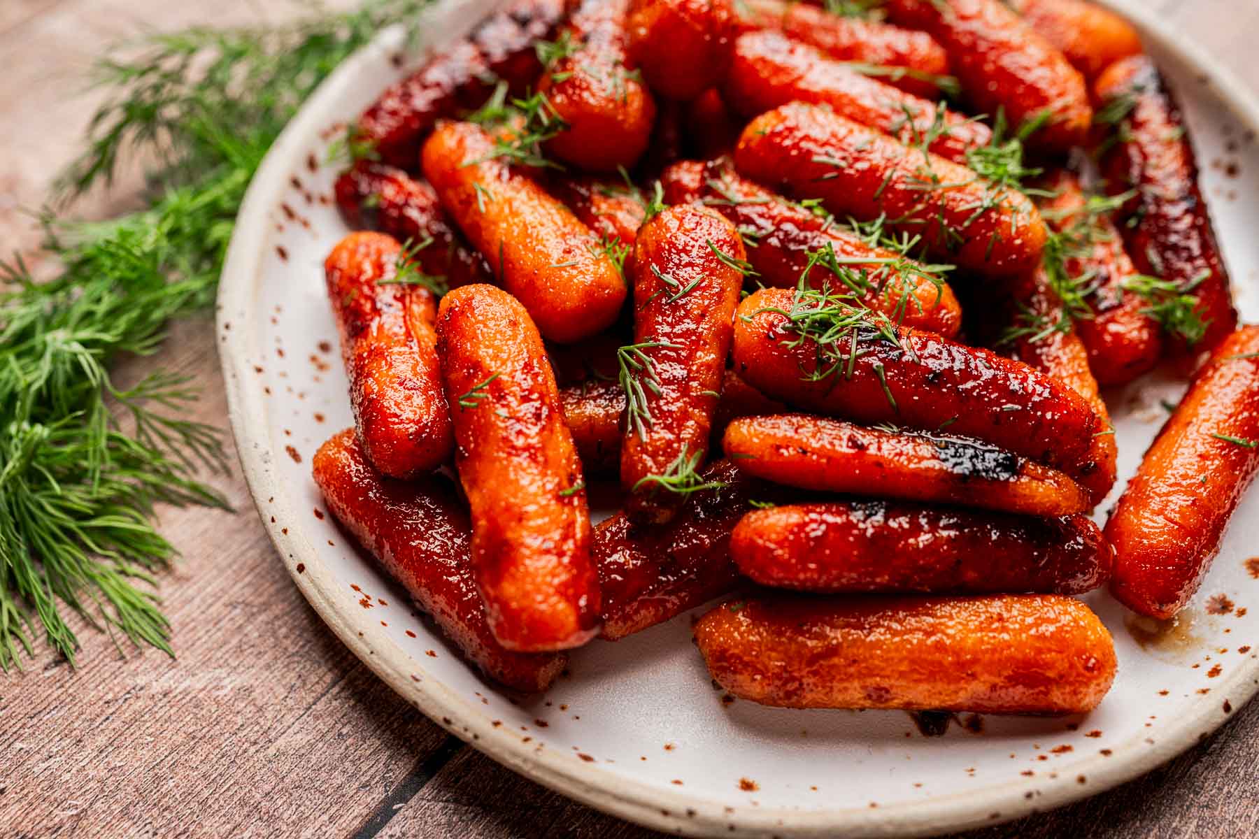A plate of brown sugar glazed carrots, roasted to perfection and garnished with fresh dill, sits on a wooden surface.