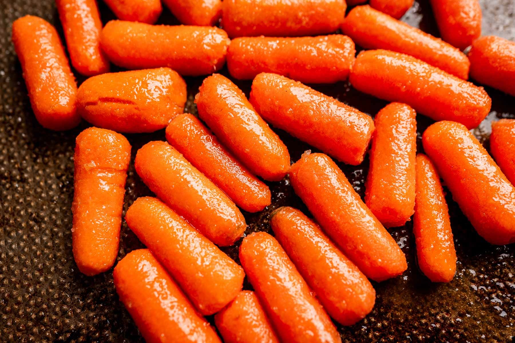 Close-up of glistening brown sugar glazed carrots spread out on a dark, textured surface.