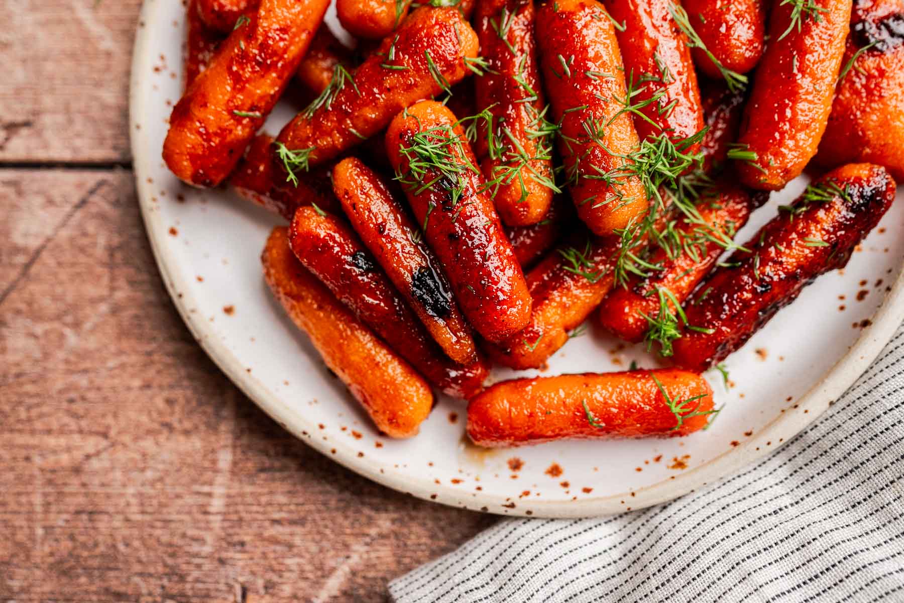 A plate of brown sugar glazed carrots garnished with fresh dill, set on a wooden table with a striped cloth nearby.