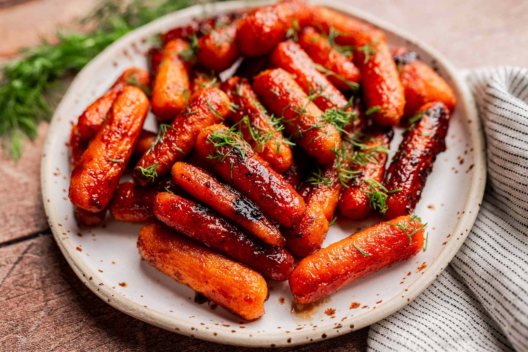 A plate of brown sugar glazed carrots garnished with fresh dill, placed on a ceramic dish next to a striped cloth napkin.