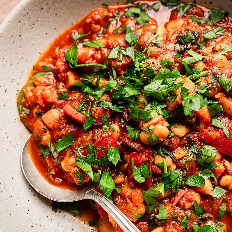 A bowl of butter bean stew in tomato sauce, filled with vegetables and garnished with chopped fresh parsley, with a spoon resting inside.
