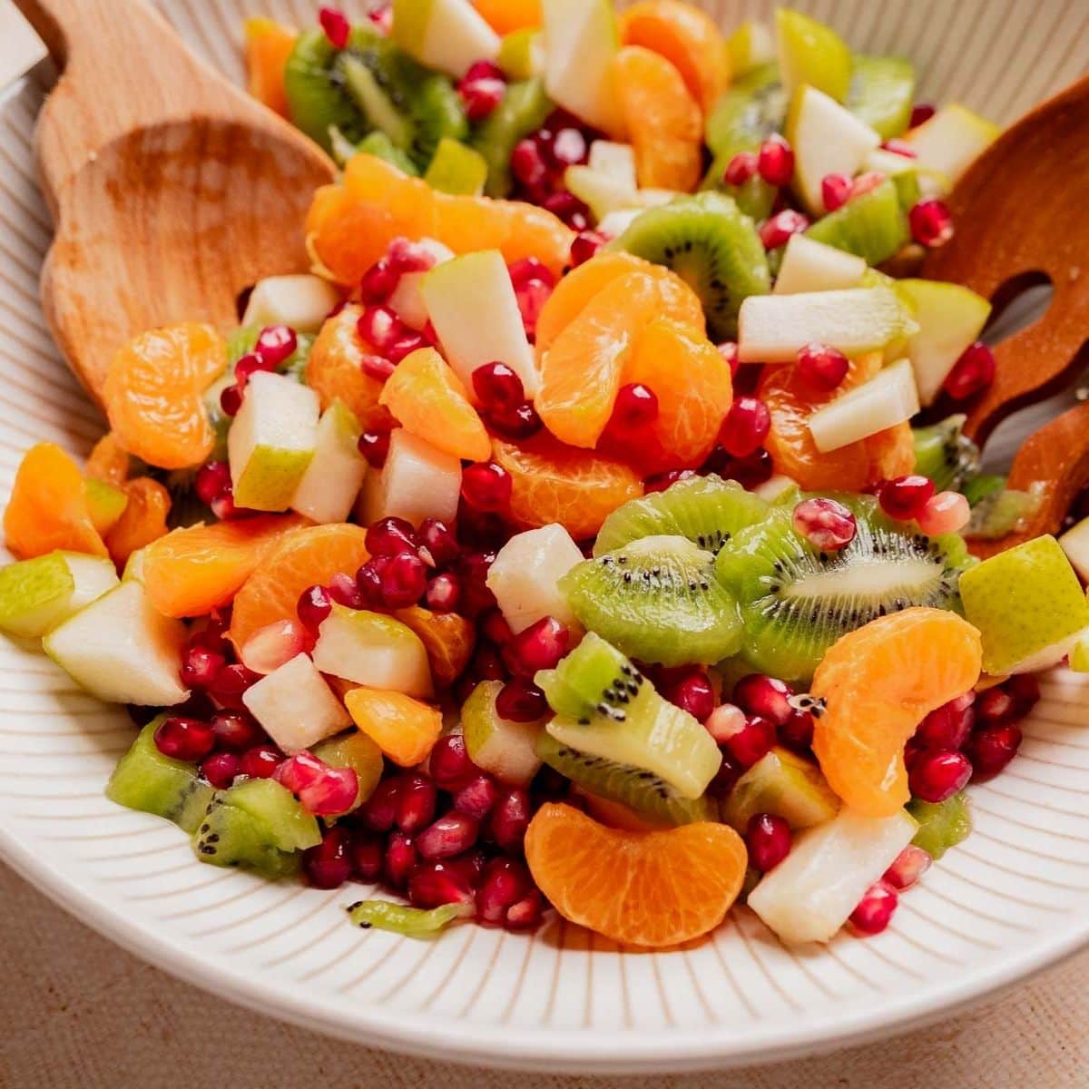 A bowl of Christmas fruit salad featuring kiwi, mandarin orange segments, pear, and pomegranate seeds, served with wooden utensils.
