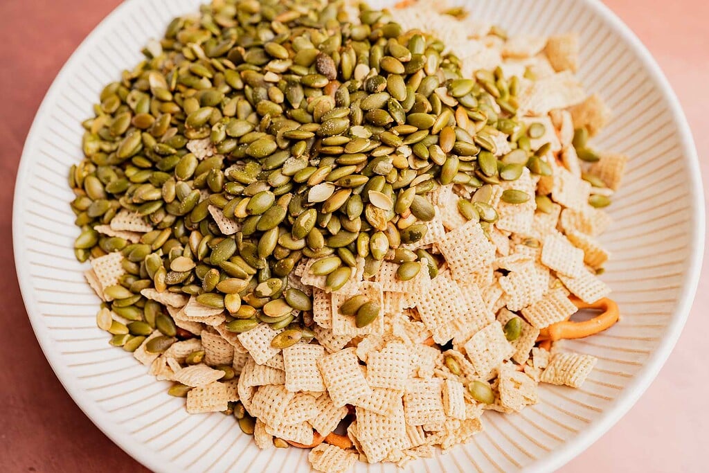 A festive bowl filled with Chex cereal, pretzel sticks, and a large amount of raw pumpkin seeds makes for a tasty Christmas snack mix.