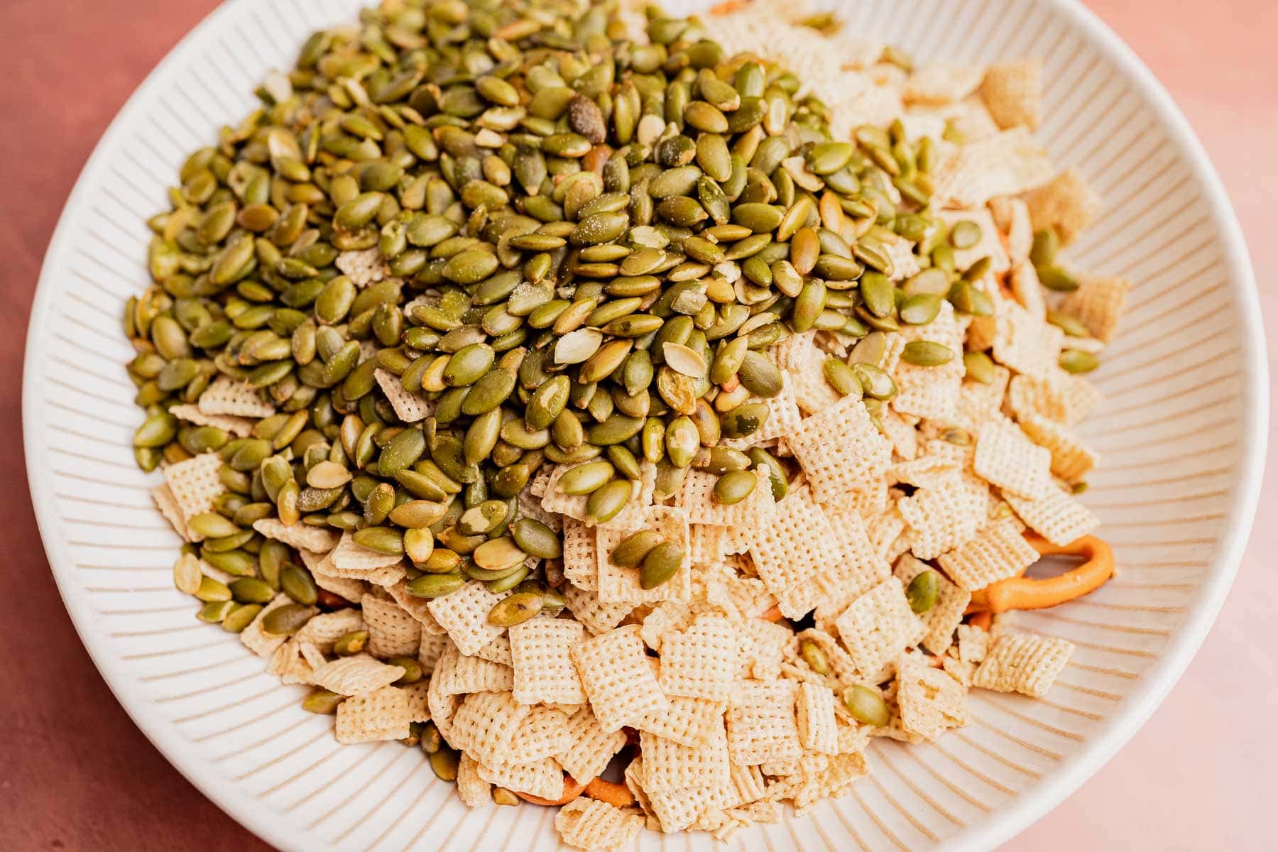 A festive bowl filled with Chex cereal, pretzel sticks, and a large amount of raw pumpkin seeds makes for a tasty Christmas snack mix.